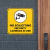 Yellow No Soliciting sign on a gray brick wall reading "Security Cameras in Use" with a security camera icon